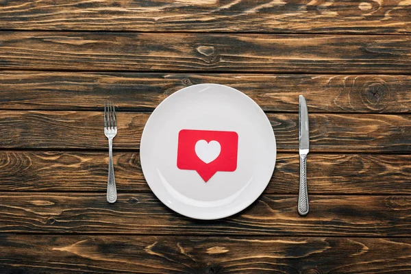 Vista superior de la placa blanca con tarjeta de corte de papel rojo con símbolo del corazón cerca del cuchillo y tenedor en la superficie de madera marrón — Stock Photo