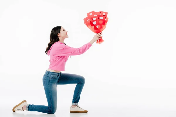 Side view of young woman standing on knee and holding bouquet of red paper cut cards with hearts symbols isolated on white — Stock Photo