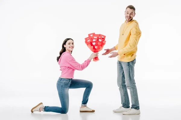 Pretty girl standing on knee and holding bouquet of red paper cut cards with hearts symbols near smiling boyfriend on white background — Stock Photo