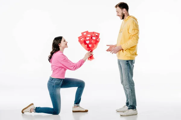 Smiling attractive girl standing on knee and holding bouquet of red paper cut cards with hearts symbols near cheerful boyfriend on white background — Stock Photo