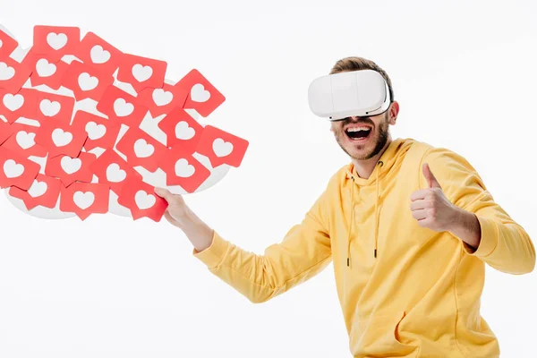 Excited man in virtual reality headset showing thumb up while holding red paper cut cards with hearts symbols isolated on white — Stock Photo