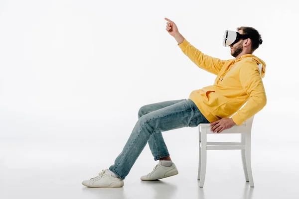 Vue latérale de l'homme en réalité virtuelle casque assis sur la chaise et pointant du doigt sur fond blanc — Photo de stock