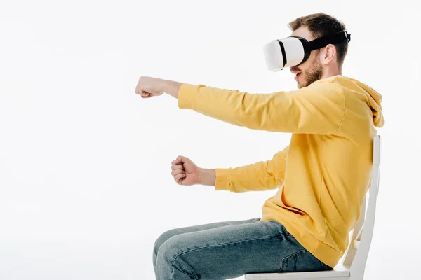 Seitenansicht eines jungen Mannes in einem Virtual-Reality-Headset, das das Autofahren imitiert, während er isoliert auf einem Stuhl auf Weiß sitzt — Stockfoto