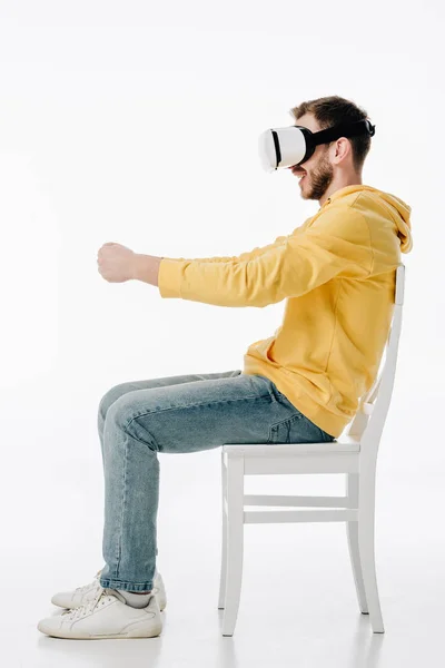 Vista lateral del joven en auriculares vr imitando la conducción mientras está sentado en la silla sobre fondo blanco - foto de stock