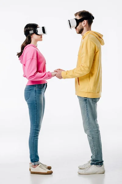 Jovem homem em mulher em jeans azuis e capuzes de mãos dadas ao usar fones de ouvido realidade virtual no fundo branco — Fotografia de Stock