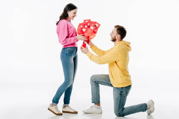 Young man standing on knee and gifting bouquet of red paper cut cards with hearts symbols to smiling girl on white background — Stock Photo