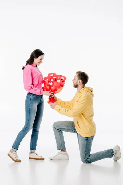 Schöner Mann, der auf Knien steht und einem hübschen Mädchen auf weißem Hintergrund einen Strauß roter Karten mit Herzsymbolen schenkt — Stockfoto