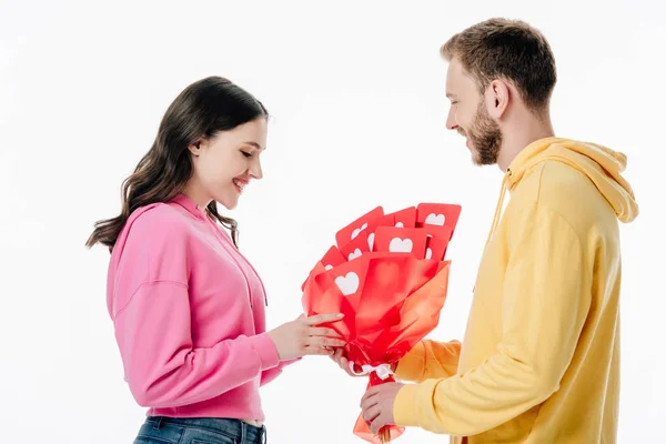 Schöner Mann, der einem hübschen lächelnden Mädchen einen Strauß roter Karten mit Herzsymbolen schenkt, isoliert auf weißem Papier — Stockfoto