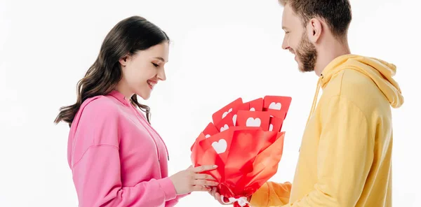 Smiling young man gifting bouquet of red paper cut cards with hearts symbols to beautiful girl isolated on white — Stock Photo