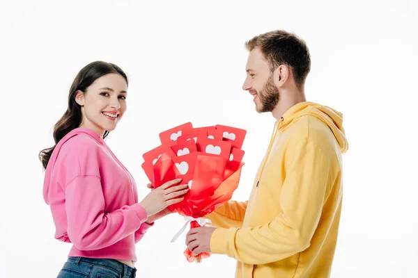 Happy young girl taking bouquet of red paper cut cards with hearts symbols from handsome boyfriend isolated on white — Stock Photo