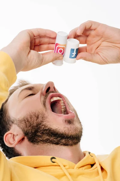 Joven barbudo hombre imitando tragar pastillas de contenedor con facebook e instagram logotipos aislados en blanco - foto de stock