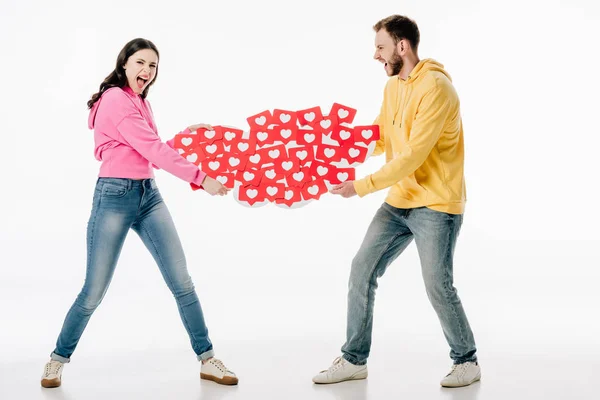 Jovem casal em jeans azuis e capuzes brigando enquanto segurando cartões de papel vermelho com símbolos de corações no fundo branco — Fotografia de Stock