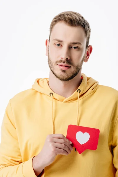 Pensativo joven sosteniendo tarjeta de corte de papel rojo con símbolo del corazón y mirando a la cámara aislada en blanco - foto de stock