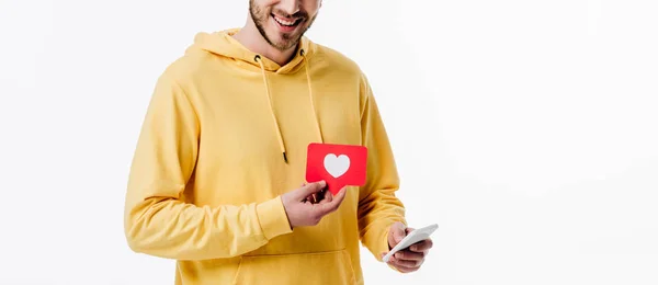 Panoramic shot of young man in yellow hoodie holding smartphone and red paper cut card with heart symbol isolated on white — Stock Photo