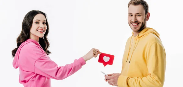 Colpo panoramico di ragazza sorridente che dà carta tagliata di carta rossa con simbolo del cuore al bel fidanzato con smartphone isolato su bianco — Foto stock