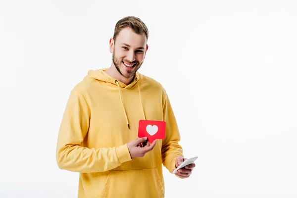 Cheerful young man using smartphone while holding red paper cut card with heart symbol isolated on white — Stock Photo