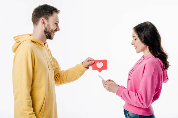 Вид збоку усміхненого молодого чоловіка, що дає червоний папір вирізати карту з символом серця подрузі, використовуючи смартфон ізольовано на білому — стокове фото