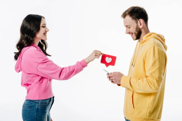 Side view of pretty girl giving red paper cut card with heart symbol to boyfriend using smartphone isolated on white — Stock Photo
