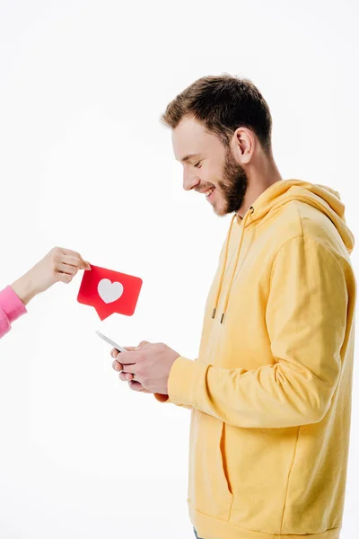 Vista recortada de la mujer que da tarjeta de corte de papel rojo con símbolo del corazón para el hombre joven utilizando un teléfono inteligente aislado en blanco - foto de stock