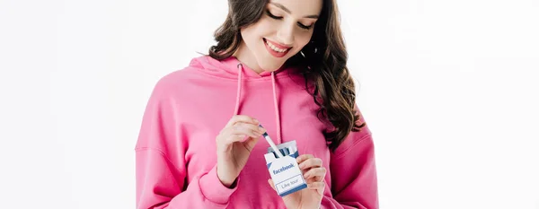 Panoramic shot of cheerful young woman holding cigarette pack with facebook logo isolated on white — Stock Photo