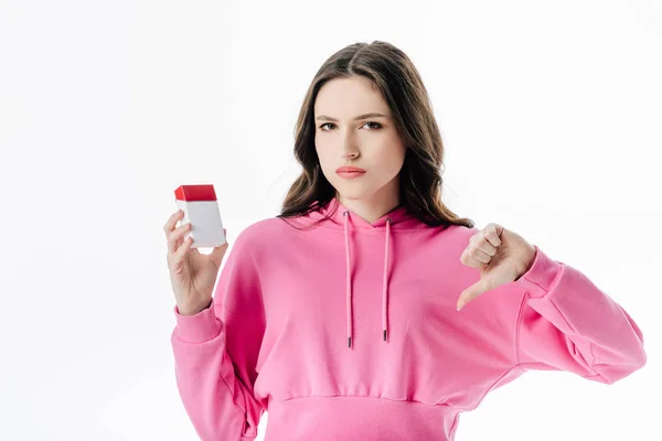 Chica seria con capucha rosa sosteniendo paquete de cigarrillos y mostrando el pulgar hacia abajo aislado en blanco - foto de stock