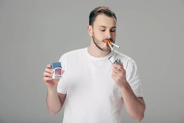 Beau jeune homme avec cigarettes dans la bouche tenant paquet avec logo instagram isolé sur gris — Photo de stock