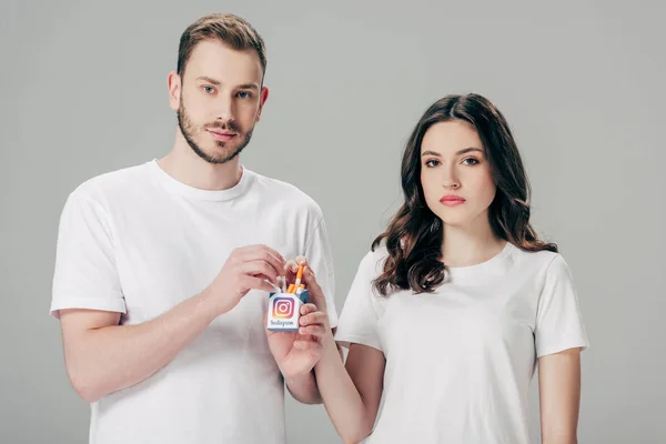 Jovem homem e mulher sério em camisetas brancas segurando maço de cigarro com logotipo instagram e olhando para a câmera isolada em cinza — Fotografia de Stock