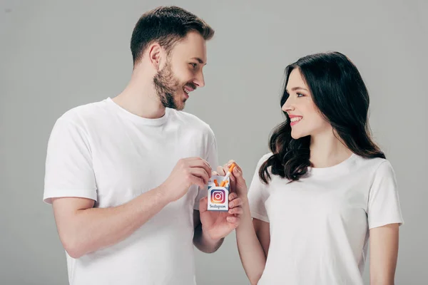 Улыбающиеся мужчина и женщина в белых футболках, держащие пачку сигарет с логотипом Instagram и глядя друг на друга изолированы на сером — стоковое фото