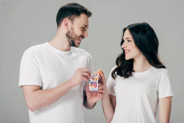 Smiling man and woman in white t-shirts holding cigarette pack with instagram logo and looking at each other isolated on grey — Stock Photo