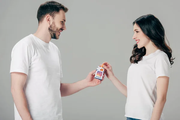 Alegre hombre y mujer en camisetas blancas sosteniendo paquete de cigarrillos con el logotipo de instagram y mirándose aislados en gris - foto de stock