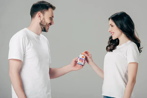 Cheerful man and woman in white t-shirts holding cigarette pack with instagram logo and looking at each other isolated on grey — Stock Photo