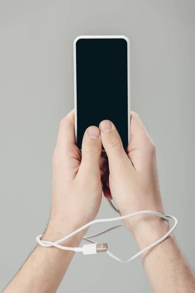 Обрізаний чоловік з usb кабелем на руках тримає смартфон з порожнім екраном ізольовано на сірому — стокове фото