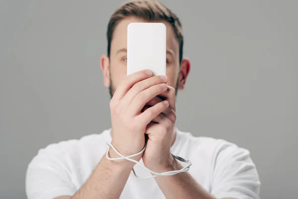 Jeune homme avec câble USB autour des mains en utilisant smartphone isolé sur gris — Photo de stock