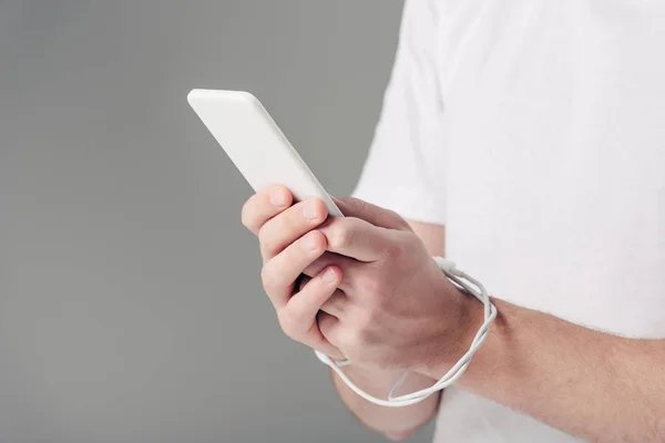 Vista cortada de jovem com cabo USB em torno de mãos usando smartphone isolado em cinza — Fotografia de Stock