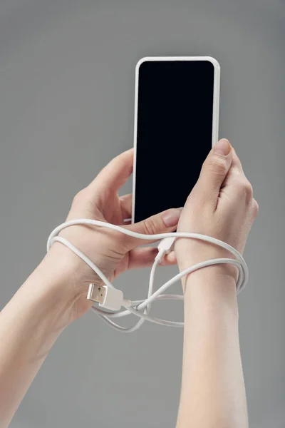Vista recortada de la mujer joven con cable USB alrededor de las manos sosteniendo teléfono inteligente con pantalla en blanco aislado en gris - foto de stock