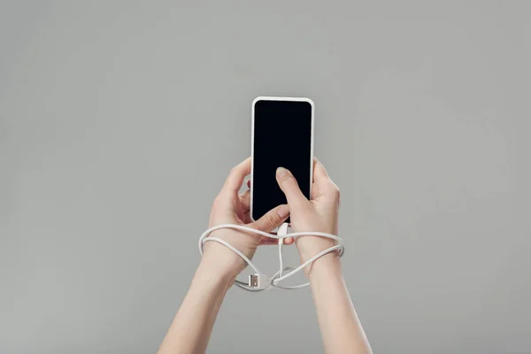 Vue partielle de la jeune femme avec câble USB autour des mains tenant smartphone avec écran blanc isolé sur gris — Photo de stock