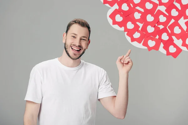 Cheerful young man in white t-shirt pointing with finger at red paper cut cards with hearts symbols isolated on grey — Stock Photo