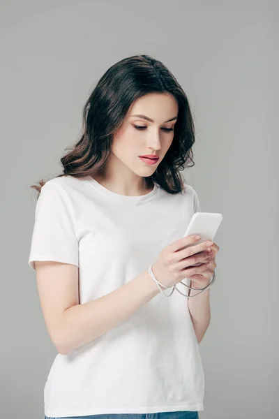 Jeune fille concentrée en t-shirt blanc en utilisant smartphone isolé sur gris — Photo de stock