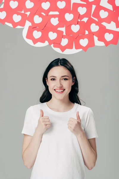 Alegre chica bonita en camiseta blanca mostrando los pulgares hacia arriba cerca de tarjetas de corte de papel rojo con corazones símbolos aislados en gris - foto de stock