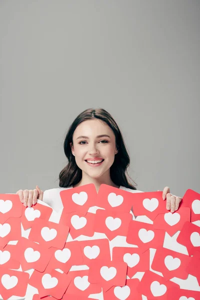 Lächelndes junges Mädchen, das rote Karten mit Herzsymbolen in der Hand hält und isoliert in die Kamera auf grau blickt — Stockfoto