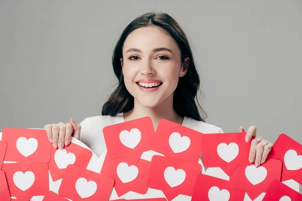 Fröhliches junges Mädchen, das rote Karten mit Herzsymbol in der Hand hält und isoliert in die Kamera auf grau blickt — Stockfoto