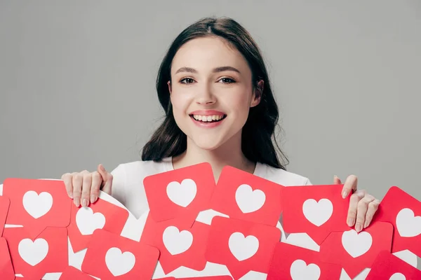 Alegre joven sosteniendo tarjetas de papel rojo cortadas con el símbolo de corazones y mirando a la cámara aislada en gris - foto de stock