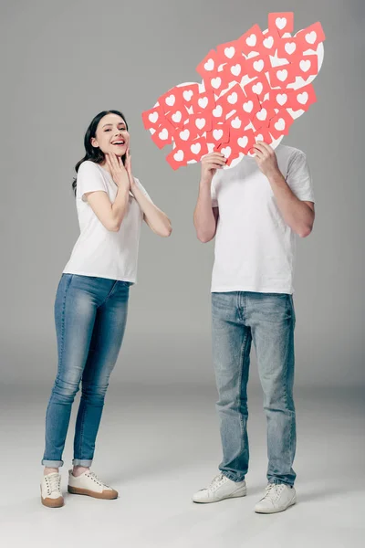 Jeune homme cachant visage derrière papier rouge cartes coupées avec des coeurs symbole près de fille excitée sur fond gris — Photo de stock