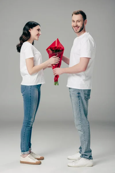 Schöner junger Mann schenkt Strauß roter Rosen an hübsche Freundin auf grauem Hintergrund — Stockfoto