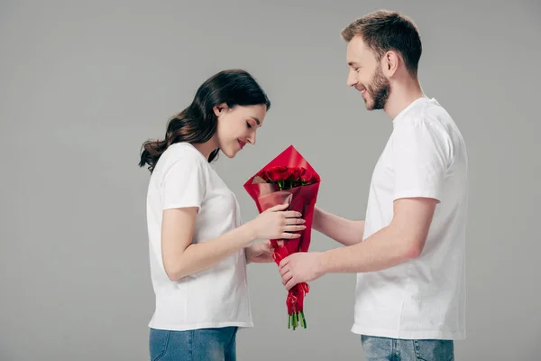 Joven sonriente hombre regalando ramo de rosas rojas a novia feliz aislado en gris - foto de stock