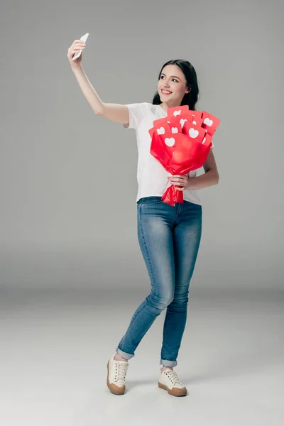 Счастливая девушка в синих джинсах держит букет красной бумаги вырезанные карточки с символом сердца и делает селфи со смартфоном на сером фоне — стоковое фото
