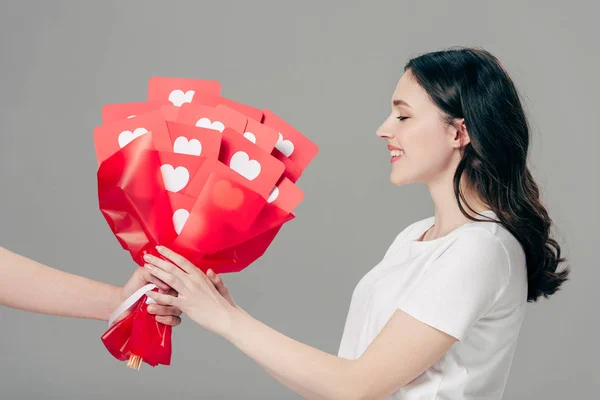Partial view of man gifting bouquet of red paper cut cards with hearts symbols to pretty girl isolated on grey — Stock Photo