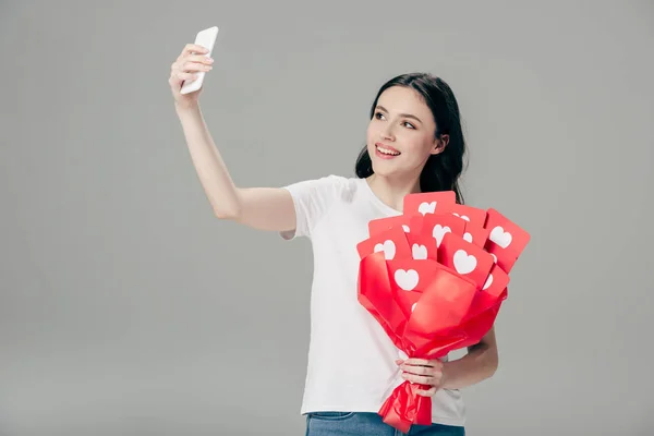 Niña sonriente sosteniendo ramo de tarjetas de corte de papel rojo con símbolo de corazones y tomar selfie con teléfono inteligente aislado en gris - foto de stock