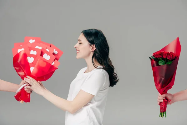 Fille souriante prenant bouquet de cartes rouges coupées en papier près de la main masculine avec bouquet de roses isolées sur gris — Photo de stock