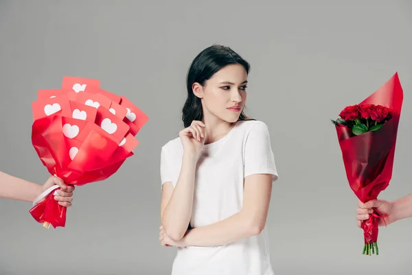 Vista recortada de manos masculinas con ramo de rosas y ramo de tarjetas de corte de papel rojo cerca de atractiva joven aislada en gris - foto de stock